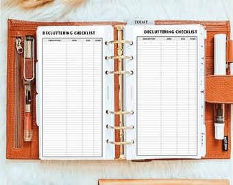 May include: A tan leather six-ring binder open to two pages titled Decluttering Checklist with columns for description, area, due, and done. The binder features a gold metal ring mechanism, a pen holder with a clear pen, and a white pen.