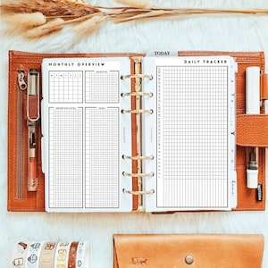 Puede incluir: Una agenda blanca con páginas de resumen mensual y seguimiento diario. La agenda tiene un encuadernador dorado y está abierta para mostrar las páginas. La agenda está sobre una superficie blanca con una cartera de cuero marrón y cinta washi.