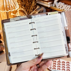 Puede incluir: Una agenda de anillas de cuero marrón abierta en una vista semanal con páginas blancas que muestran texto negro para los días de la semana, un calendario mensual y secciones de cuadrícula. Un bolígrafo plateado está en el lazo lateral.