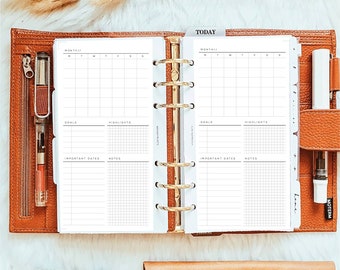 Puede incluir: Una agenda de cuero color canela con seis anillas abierta en dos páginas blancas con una cuadrícula de calendario mensual, secciones para objetivos, destacados, fechas importantes y notas. Incluye un mecanismo de anillas dorado y un bolígrafo.