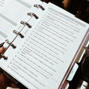 May include: Open planner with a car maintenance checklist and a self-care list. The car checklist includes service items like oil level and hoses. The self-care list features activities like exercise, healthy eating, and journaling. The planner has metal rings and labeled tabs.