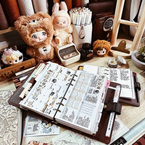 May include: A brown leather ring-bound planner open to a weekly spread with decorative stickers, surrounded by plush bear and rabbit figures, a white pen holder with markers, a small ink bottle, and a black cutting mat with 30x90 cm text.