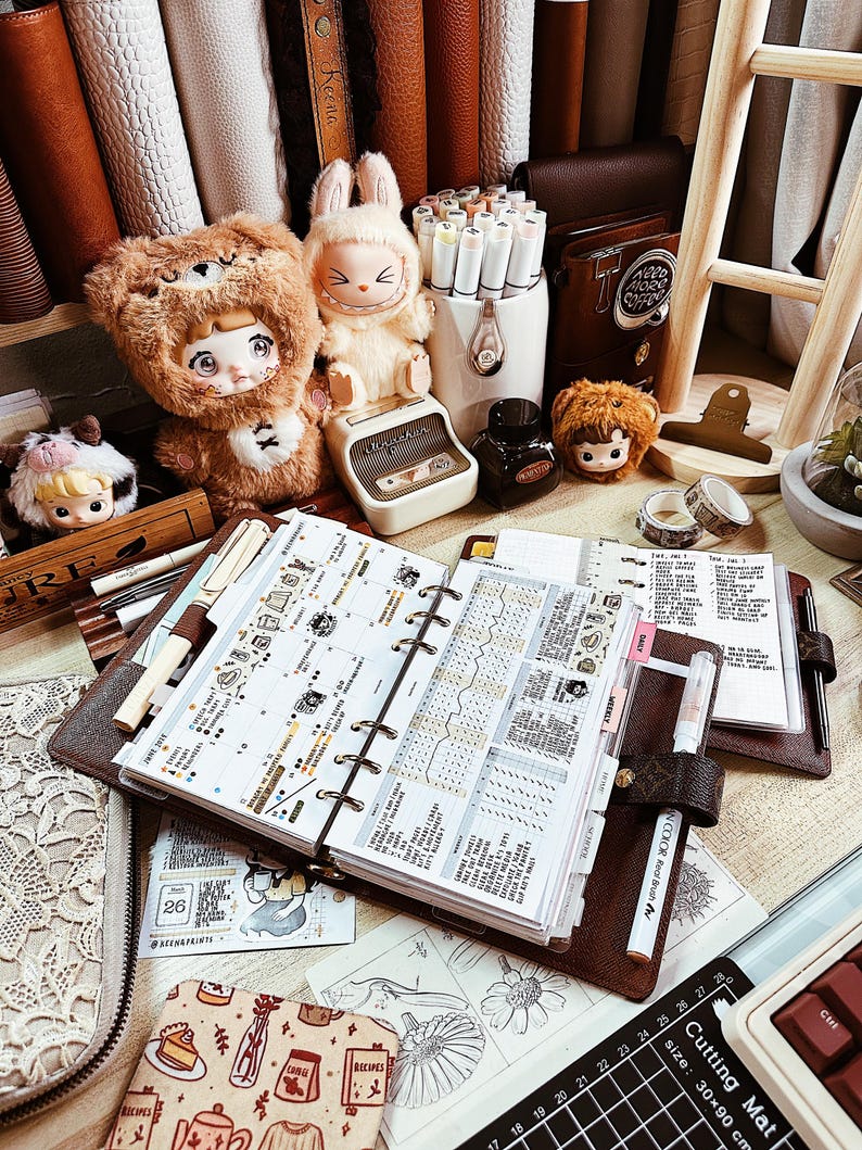 May include: A brown leather ring-bound planner open to a weekly spread with decorative stickers, surrounded by plush bear and rabbit figures, a white pen holder with markers, a small ink bottle, and a black cutting mat with 30x90 cm text.