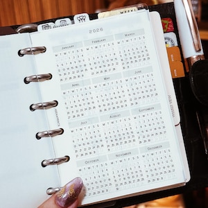 Puede incluir: Una agenda personal con un mecanismo de encuadernación de seis anillas que contiene un calendario anual para 2026. El papel blanco presenta un diseño de cuadrícula para los doce meses, con el texto 2026, e incluye un bolígrafo blanco con detalles dorados.