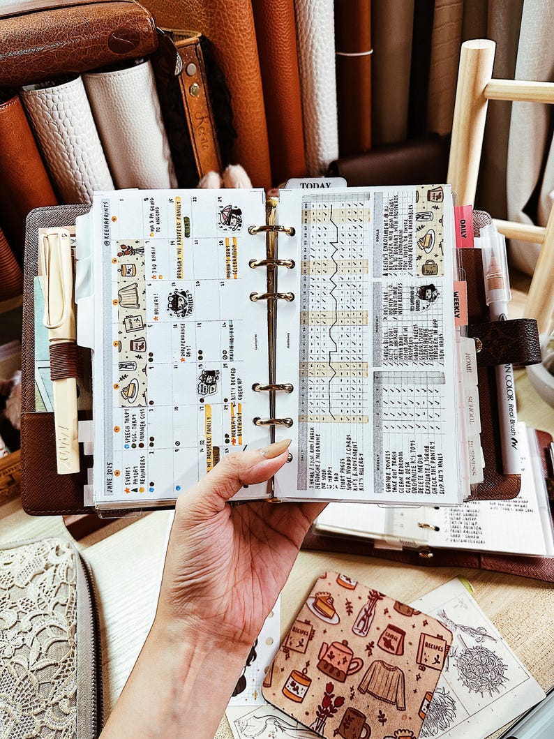 May include: A brown leather personal planner with a six-ring metal binder mechanism, open to a monthly calendar page and a habit tracker page. The planner includes a cream-colored pen, a white pen, and various decorative stickers with illustrations.
