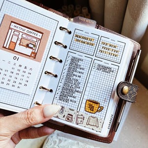 May include: A brown leather personal planner with gold rings, featuring a grid-paper insert with a Meow Coffee illustration, a calendar for the month of January, and a task list titled This Week with handwritten food goals and a yellow mug sticker.