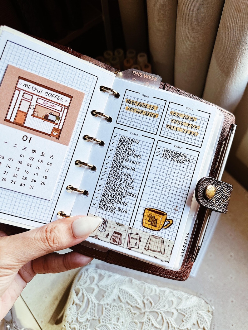 May include: A brown leather personal planner with gold rings, featuring a grid-paper insert with a Meow Coffee illustration, a calendar for the month of January, and a task list titled This Week with handwritten food goals and a yellow mug sticker.