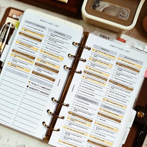 May include: An open planner with a "Gratitude List" layout, featuring sections like "Moments to Remember" and "Today." Gold-toned rings bind the pages. A vintage-style radio and pens are visible in the background.