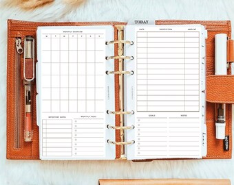 Puede incluir: Una agenda de cuero marrón con seis anillas abierta mostrando páginas blancas con una cuadrícula de resumen mensual, secciones para fechas importantes, tareas, una tabla financiera, objetivos y notas, junto a un bolígrafo y cintas decorativas.