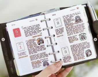 May include: A personal planner with a dark brown leather cover and silver ring binding. The open pages feature a grid layout with handwritten notes, dates for May, and small cartoon character stickers. A silver pen is attached to the side.