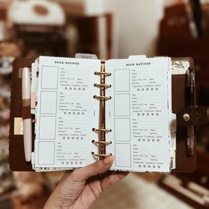 May include: A brown leather ring-bound planner featuring white pages with printed book rating templates. Each page includes fields for title, author, genre, series, pages, dates, and a five-star rating system. A light pink pen and a black pen are attached.