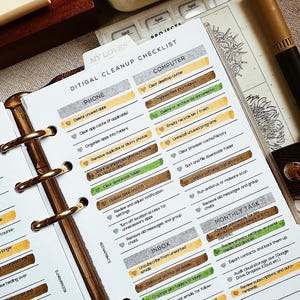May include: Planner open to a digital cleanup checklist. The checklist includes tasks for phone, computer, inbox, and monthly tasks. The planner has gold rings and a brown cover. A pen and stationery items are visible.