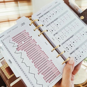 May include: A close-up of a planner with a gold binder. The planner has a habit tracker with a sleep tracker on the left page. The habit tracker has a grid with dates and days of the week. The sleep tracker has a graph with a line showing the amount of sleep each night. The habit tracker has a list of habits to track, including "No Spend - Stationery", "1 Cup of Coffee Per Day", "No Sugar", "Listen to Autism Podcast", and "Workout / Walk".