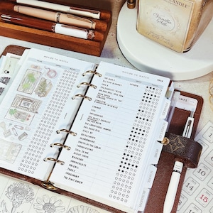 May include: A brown leather ring-bound planner open to a Movies to Watch page with a list of horror film titles and star ratings. The planner features tabbed dividers labeled Finances, Schedules, Home, School, and Business, with a white pen attached.