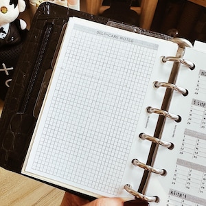 May include: A black leather planner with a grid-lined page labeled "SELF-CARE NOTES" is open, revealing a calendar. The planner has silver metal rings and a zipper closure. The page is held open by a hand with dark red nail polish.