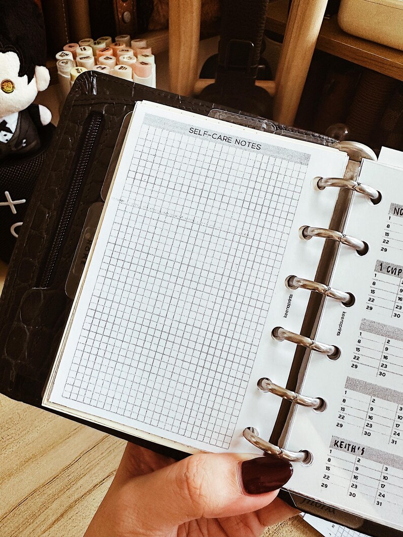 May include: A black leather planner with a grid-lined page labeled "SELF-CARE NOTES" is open, revealing a calendar. The planner has silver metal rings and a zipper closure. The page is held open by a hand with dark red nail polish.