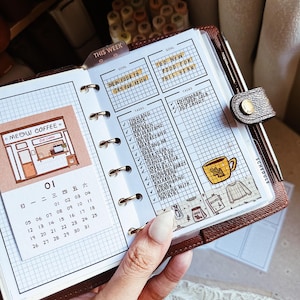 May include: A brown leather ring-bound planner featuring a grid-paper insert with a calendar for the month of 01, a Meow Coffee shop illustration, and a task list with handwritten entries including Tinolang Manok and Pork Steak.