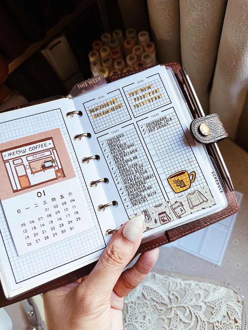 May include: A brown leather ring-bound planner featuring a grid-paper insert with a calendar for the month of 01, a Meow Coffee shop illustration, and a task list with handwritten entries including Tinolang Manok and Pork Steak.