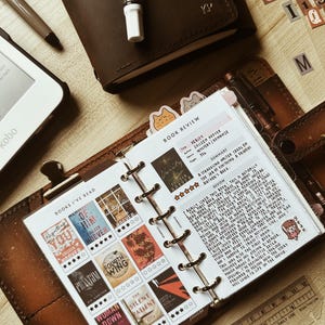 May include: A personal planner open to a page titled Books I've Read and Book Review, featuring miniature book cover stickers, star ratings, and handwritten notes. The planner includes a metal ring binder, a brown leather cover, and a clear plastic ruler.