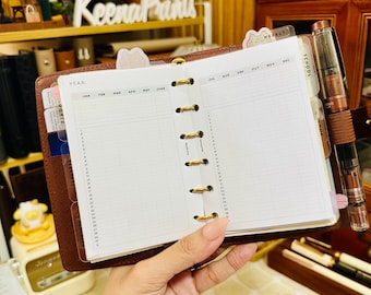 Può includere: Un'agenda ad anelli in pelle marrone con inserti in carta bianca dotati di griglia per il calendario annuale, intestazioni mensili da gennaio a dicembre e righe giornaliere numerate. L'agenda include anelli in metallo dorato e una penna stilografica.