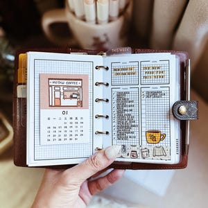 May include: A brown leather ring-bound planner open to a page with a grid layout. The left side features a Meow Coffee illustration and a calendar for the month of January. The right side displays task lists, a goal section, and a yellow coffee mug sticker.