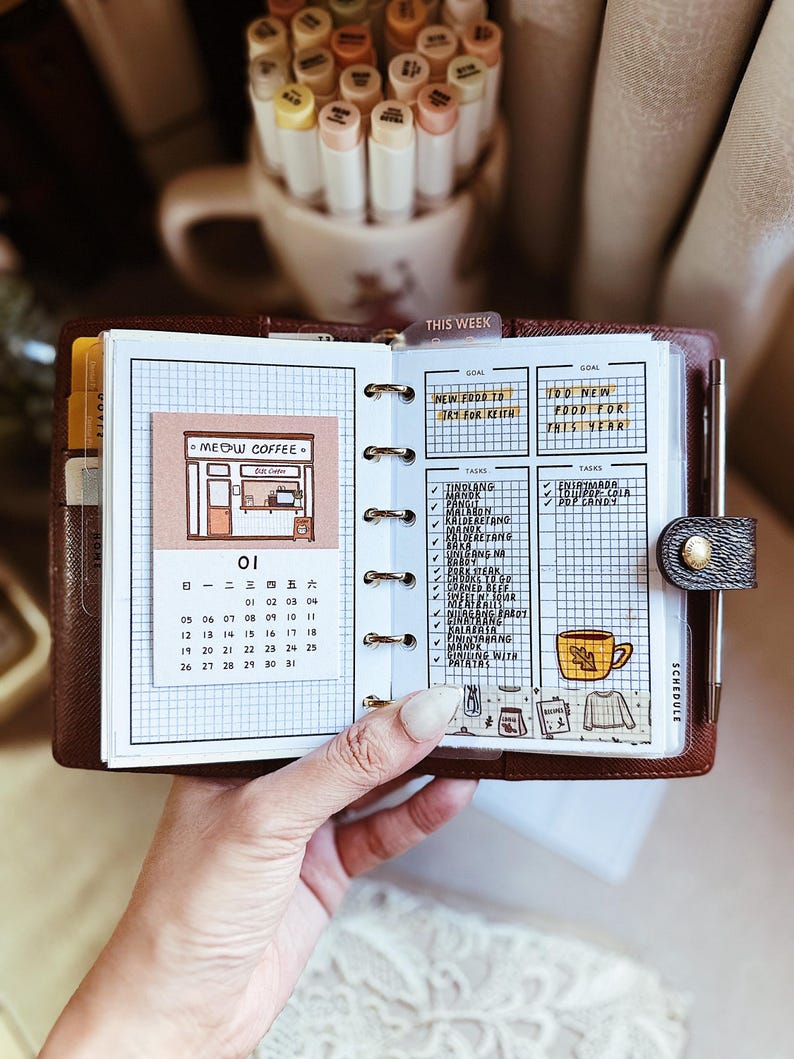 May include: A brown leather ring-bound planner open to a page with a grid layout. The left side features a Meow Coffee illustration and a calendar for the month of January. The right side displays task lists, a goal section, and a yellow coffee mug sticker.