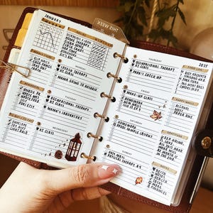 Könnte beinhalten: Ein Nahaufnahme eines braunen Lederplaners mit einem Wochenplan und einer To-Do-Liste. Der Planer ist für Januar geöffnet und enthält tägliche Aufgaben, Erinnerungen und ein Essenstagebuch. Der Planer ist mit Aufklebern und einer kleinen Laterne verziert.