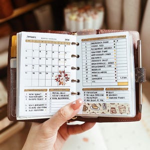 Op de afbeelding: Een kleine, witte planner met een maandelijkse kalender voor januari 2025. De planner heeft een sectie voor belangrijke data, maandelijkse taken en een sectie voor het bijhouden van financiën. De planner is open op een pagina met een lijst van financiële doelen en een sectie voor notities.