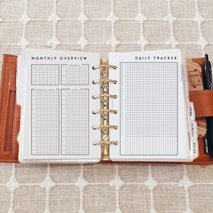 Op de afbeelding: Een bruine lederen planner met zes ringen, opengeklapt om witte pagina's met zwarte rasterindelingen te tonen. De linkerpagina is getiteld Monthly Overview met secties voor prioriteiten, data en taken. De rechterpagina is een Daily Tracker met een genummerd raster.