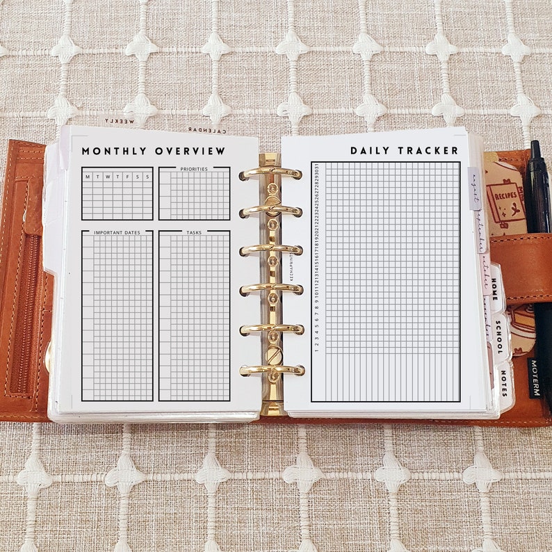 Op de afbeelding: Een bruine lederen planner met zes ringen, opengeklapt om witte pagina's met zwarte rasterindelingen te tonen. De linkerpagina is getiteld Monthly Overview met secties voor prioriteiten, data en taken. De rechterpagina is een Daily Tracker met een genummerd raster.