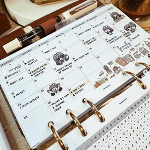 May include: A white paper planner insert for January 2024 featuring a grid layout with handwritten notes, black ink text, and small cartoon character stickers. The planner page is held by gold-colored metal rings and includes a white fountain pen.