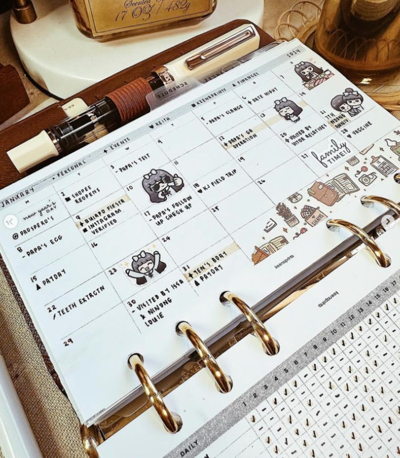 May include: A white paper planner insert for January 2024 featuring a grid layout with handwritten notes, black ink text, and small cartoon character stickers. The planner page is held by gold-colored metal rings and includes a white fountain pen.