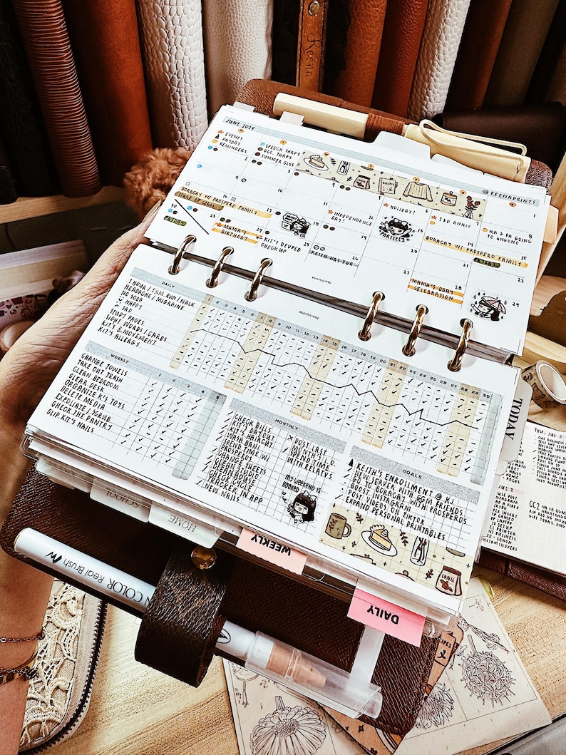 May include: A brown leather ring-bound planner open to a June 2023 calendar page. The planner features handwritten daily and weekly habit trackers, goal lists, and decorative stickers. A white pen is tucked into the leather pen loop on the cover.