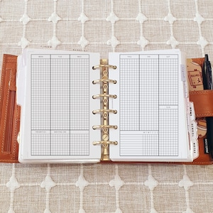 May include: Open planner with weekly schedule pages. The pages are white with black grid lines and headings for each day of the week. The planner is in a brown leather binder with gold-colored rings and a pen.