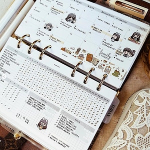 May include: A ring-bound planner open to a January 2024 calendar page with handwritten notes, small cartoon character stickers, and decorative washi tape. The lower section features a daily habit tracker grid and a list of goals with checkmarks.