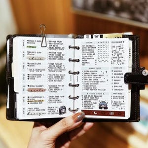 Puede incluir: Agenda abierto con notas manuscritas y un clip metálico. El planificador está abierto a una página con secciones diarias, semanales y de registro de alimentos. El texto incluye "ideas", "crear" y "diseño". El planificador es negro con cierre a presión.