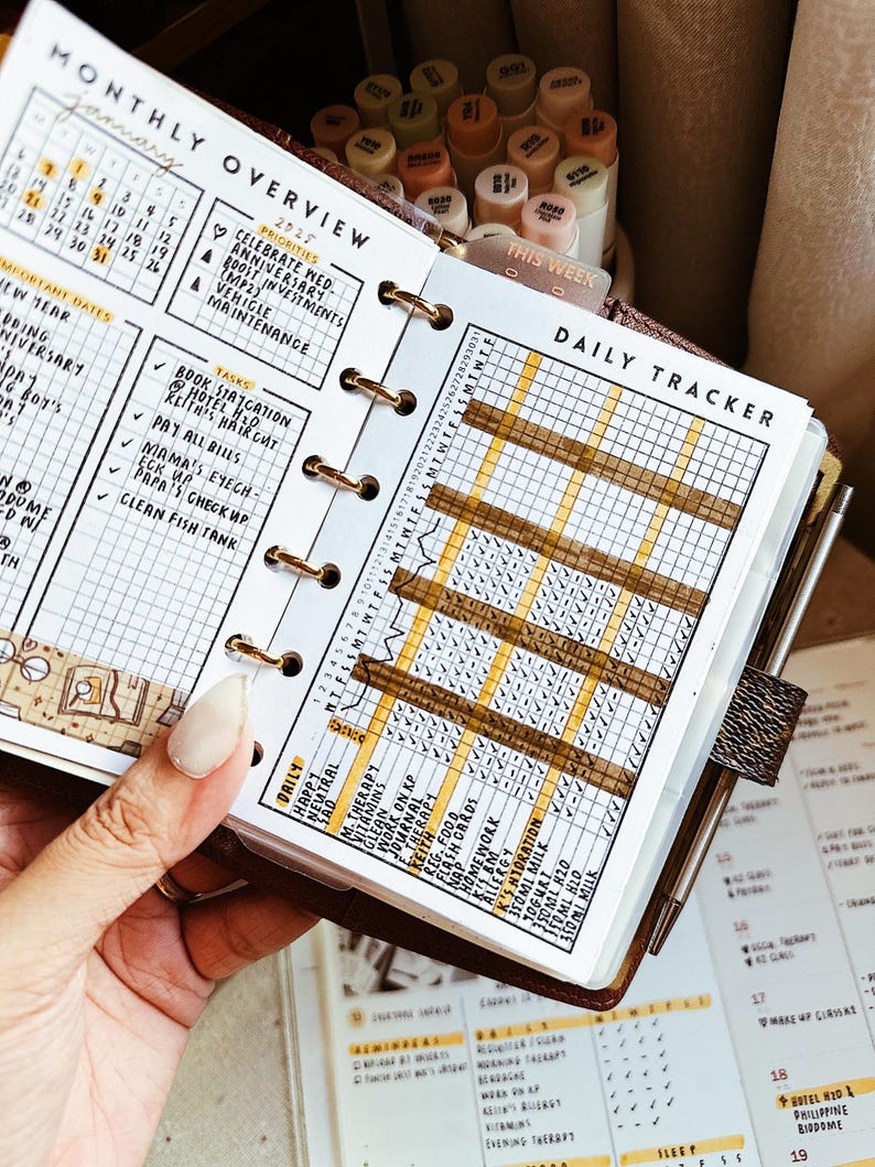 Op de afbeelding: Een persoonlijke planner open op een maandoverzicht en dagelijkse tracker pagina. Het witte papier heeft zwarte rasterlijnen, gedrukte tekst waaronder Monthly Overview, Daily Tracker, en handgeschreven taken. Gouden ringen binden de pagina's in een bruine kaft.