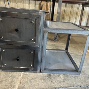May include: A dark grey metal bedside table with two drawers and a side shelf. The drawers have black knobs and are framed with a darker metal. The table is made of welded steel and has a modern, industrial design.