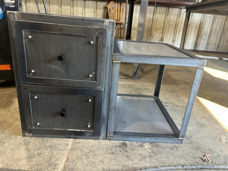 May include: A dark grey metal bedside table with two drawers and a side shelf. The drawers have black knobs and are framed with a darker metal. The table is made of welded steel and has a modern, industrial design.