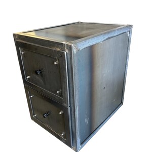May include: A two-drawer metal cabinet with a welded frame. The cabinet has a dark, industrial finish with visible weld marks. Each drawer features a black knob and is secured with screws. The overall design is boxy and modern.
