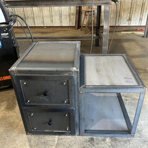 May include: A welded metal nightstand with two drawers and a side table. The nightstand has a dark, industrial finish with black knobs on the drawers. The side table has an open shelf. The metal frame is visible.
