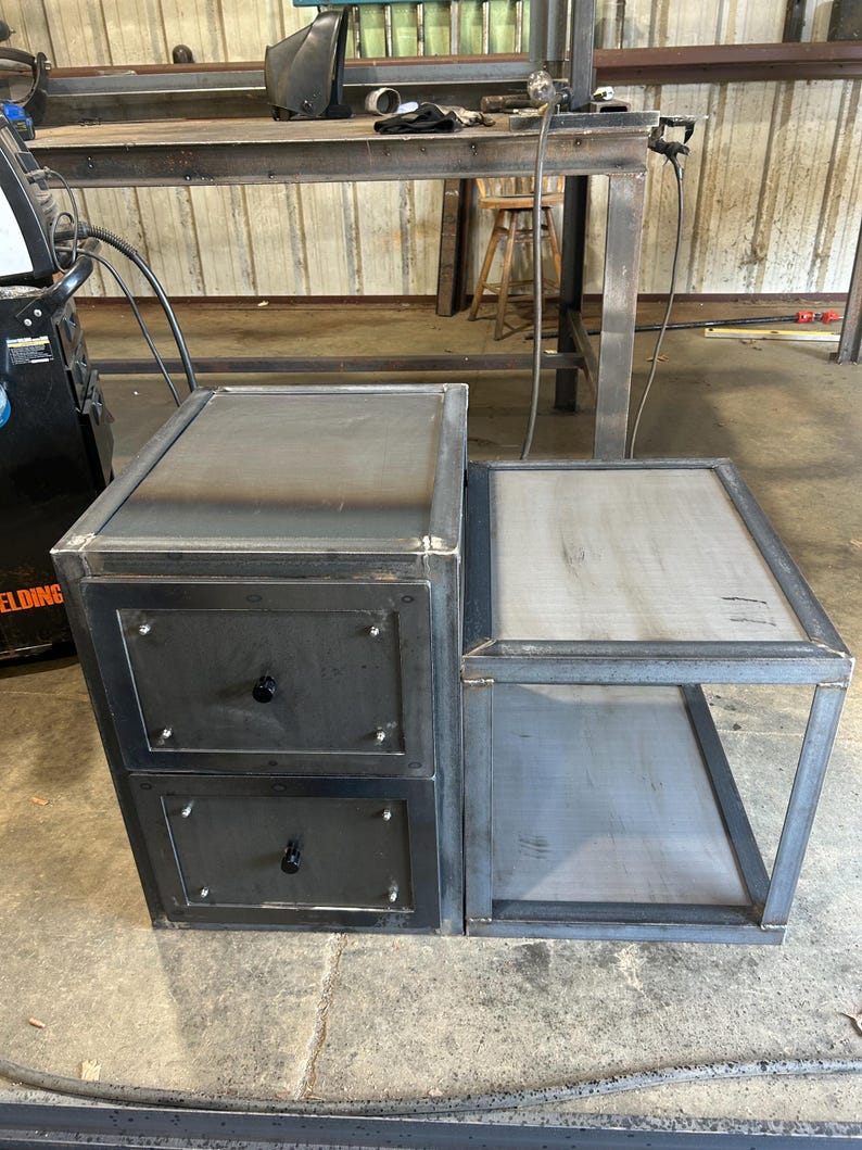 May include: A welded metal nightstand with two drawers and a side table. The nightstand has a dark, industrial finish with black knobs on the drawers. The side table has an open shelf. The metal frame is visible.