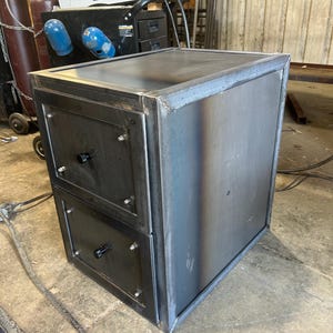 May include: A metal cabinet with two drawers and a flat top, showing visible weld marks. The cabinet is made of welded steel with a dark, industrial finish. Black drawer pulls and screws add detail. Dimensions are approximately 60cm x 60cm x 60cm.