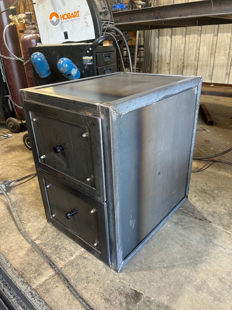 May include: A metal cabinet with two drawers and a flat top, showing visible weld marks. The cabinet is made of welded steel with a dark, industrial finish. Black drawer pulls and screws add detail. Dimensions are approximately 60cm x 60cm x 60cm.