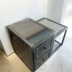 May include: A two-piece industrial-style side table set. The set features a dark grey metal frame with a rectangular top and two drawers. The drawers have black knobs and are mounted on a larger rectangular base.
