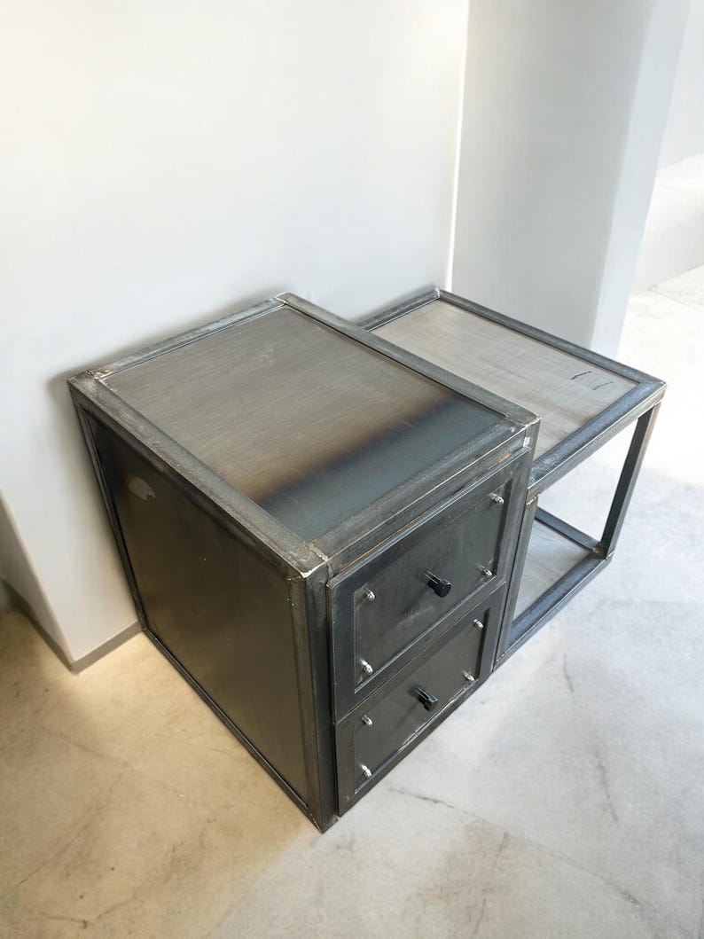 May include: A two-piece industrial-style side table set. The set features a dark grey metal frame with a rectangular top and two drawers. The drawers have black knobs and are mounted on a larger rectangular base.
