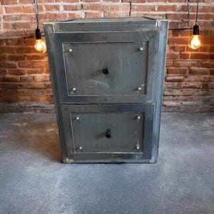 May include: A two-drawer metal cabinet in dark grey. Each drawer features a black knob and is framed by a darker metal border. The cabinet is set against a brick wall and a concrete floor.