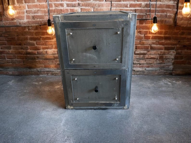 May include: A two-drawer metal cabinet in dark grey. Each drawer features a black knob and is framed by a darker metal border. The cabinet is set against a brick wall and a concrete floor.