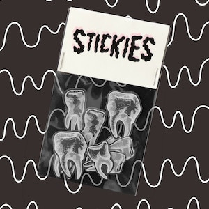 May include: A clear plastic bag with the word "STICKIES" printed on the front. The bag contains a set of black and white tooth-shaped stickers.
