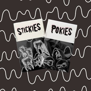 May include: Two clear plastic bags filled with enamel pins. One bag has a white label with the text "STICKIES" in black. The other bag has a white label with the text "POKIES" in black. The pins in the first bag are white tooth shapes. The pins in the second bag are black and feature a tooth shape.